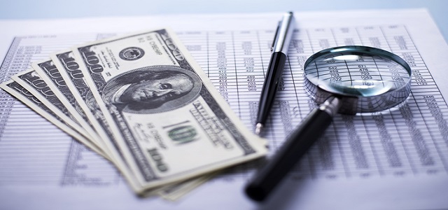 Conceptual image of fanned dollar banknotes with a magnifying glass, pen and analytical financial report lying on a table with shallow dof